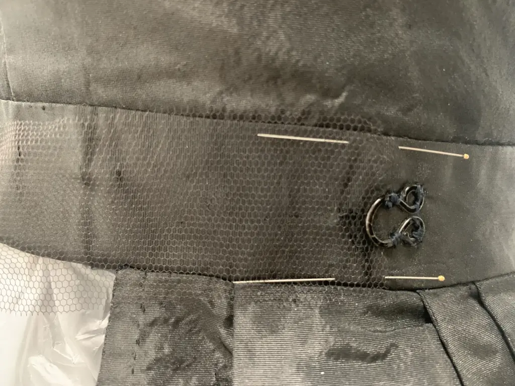 Restauración de ropa antigua: cinturilla de una prenda de seda negra cubierta con tul de soporte sujeto con alfileres, junto a un corchete metálico original.