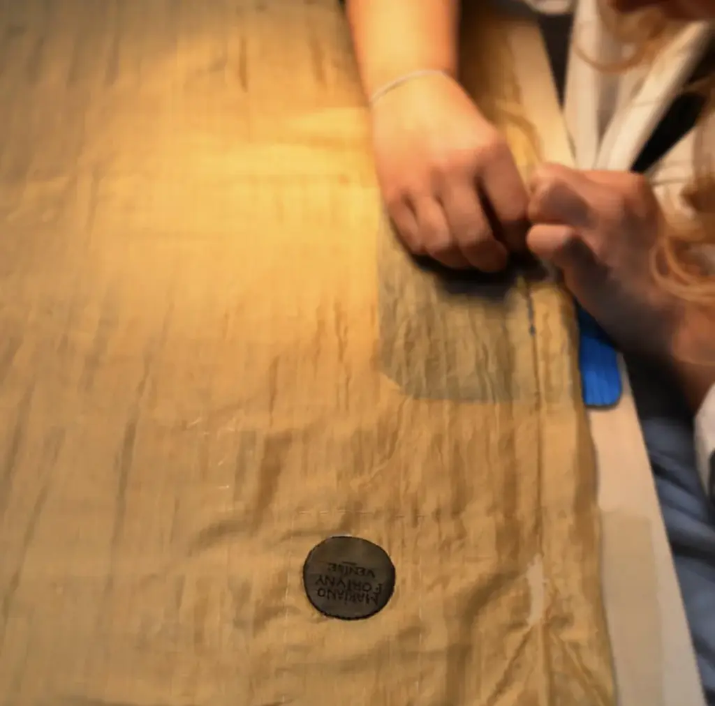 Restauración de ropa antigua: dos manos manipulan el borde de un vestido de seda plisada color oro extendido sobre la mesa de trabajo, con la etiqueta circular estampada «Mariano Fortuny Venise».