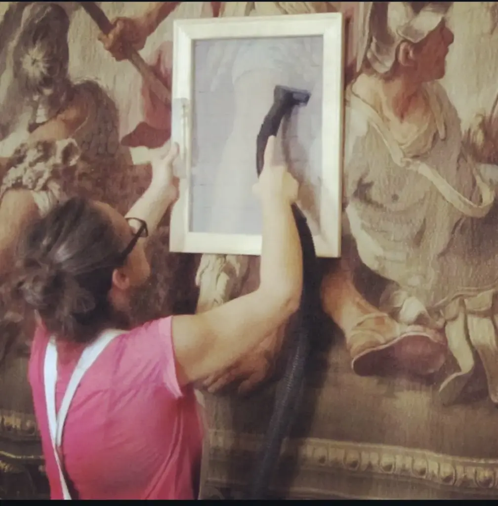 Restauración de tapices: una restauradora apoya un bastidor con malla sobre un tapiz con escena de soldados y aspira la superficie a través de la malla.